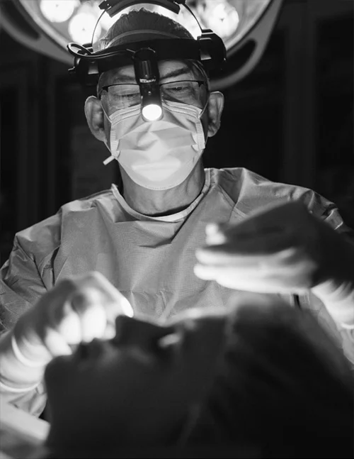 A black and white image showing Dr. Washal, a surgeon, performing a procedure on a patient. He is wearing a surgical mask, glasses, a headlamp, and surgical gloves, with his hands near the patient's face.