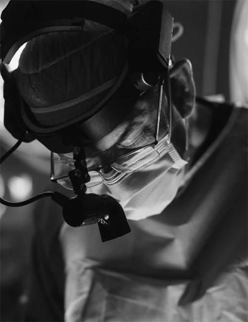 A black and white close-up image of Dr. Washal, a surgeon, focused on a procedure. He is wearing a surgical mask, glasses, a cap, and surgical loupes with an attached headlamp.