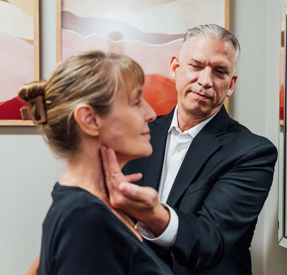 Dr. Lee in a black suit and white shirt consulting with a female patient. The patient is touching her cheek while the doctor looks toward the camera with a slight smile. A mirror and abstract artwork are visible in the background.