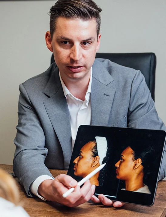 A male doctor, Dr. Washal, wearing a gray suit jacket and white shirt, is seated at a wooden desk while consulting with a female patient. He is holding a tablet displaying before and after images of another patient's facial procedure, pointing to the images with a stylus. - Alar Base Rhinoplasty in Severna Park, MD