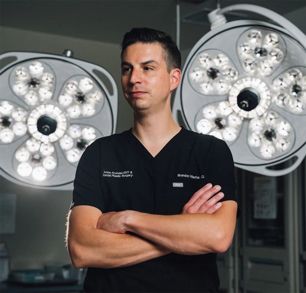 A portrait of Dr. Washal, a male doctor, standing with his arms crossed while wearing black scrubs. Two bright surgical lights are visible in the background on either side of him.