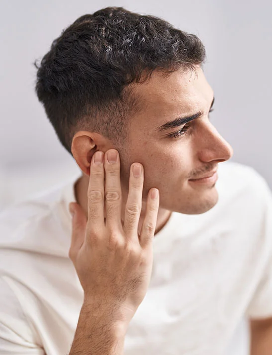 A young man with short, dark hair looks to the side, his hand lightly touching his ear. He is wearing a white t-shirt and has a slight smile. The background is a clean white with subtle grey shadows. - Ear Pinning Severna Park, MD