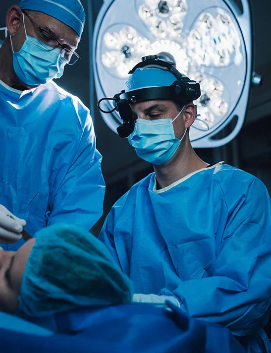 Close up of two surgeons, Dr. Washal (left) and Dr. Lee (right), wearing light blue surgical scrubs, masks, caps, and gloves, perform a reconstructive procedure on a patient lying face-up on an operating table. Dr. Lee is also wearing a surgical headlamp. The operating room is dimly lit except for the intense, focused illumination from the large overhead surgical lights directly above the patient. - Earlobe Repair in Severna Park, MD