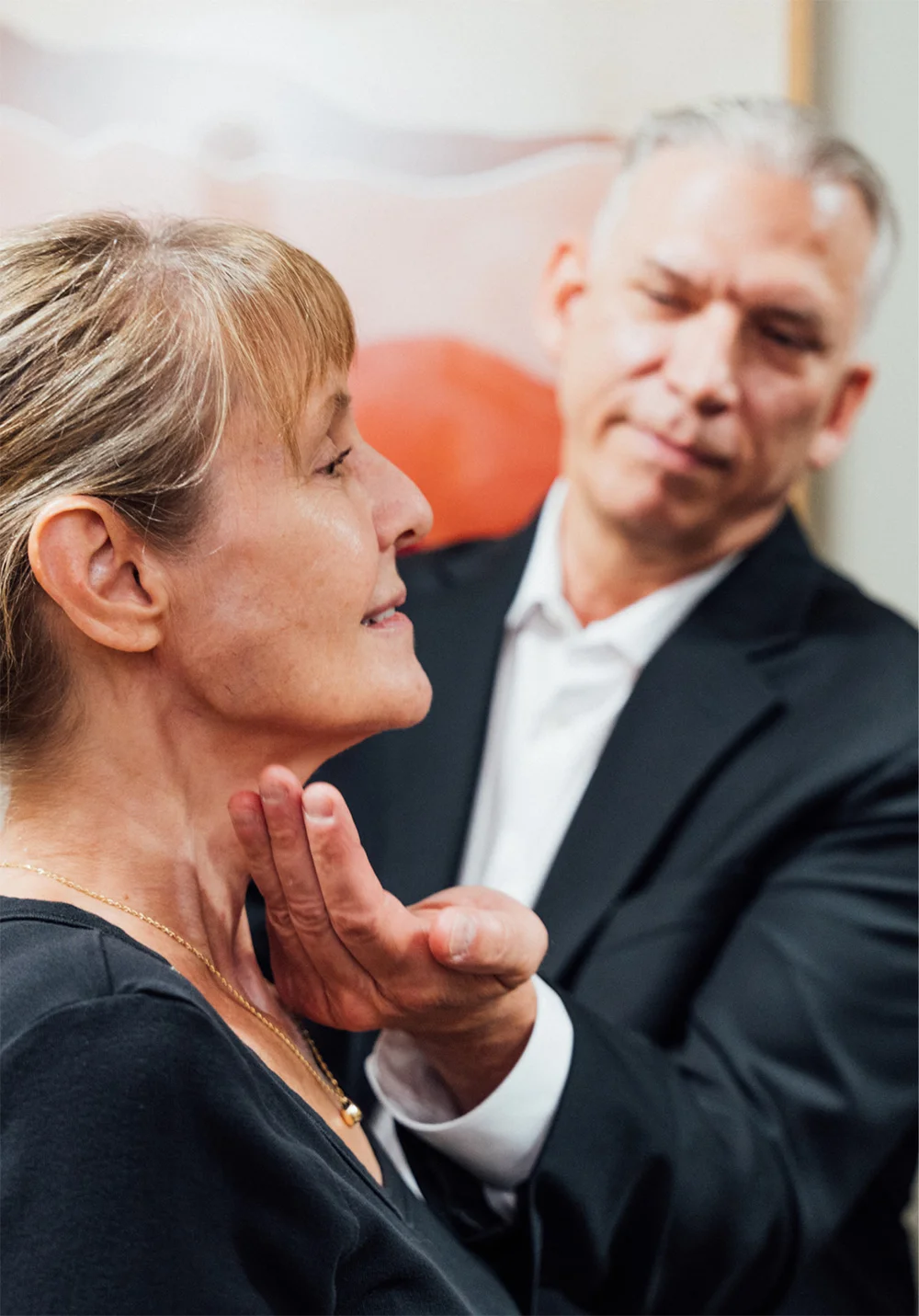 Dr. Lee in a black suit and white shirt consulting with a female patient. The patient is touching her cheek while the doctor looks toward the camera with a slight smile. A mirror and abstract artwork are visible in the background.