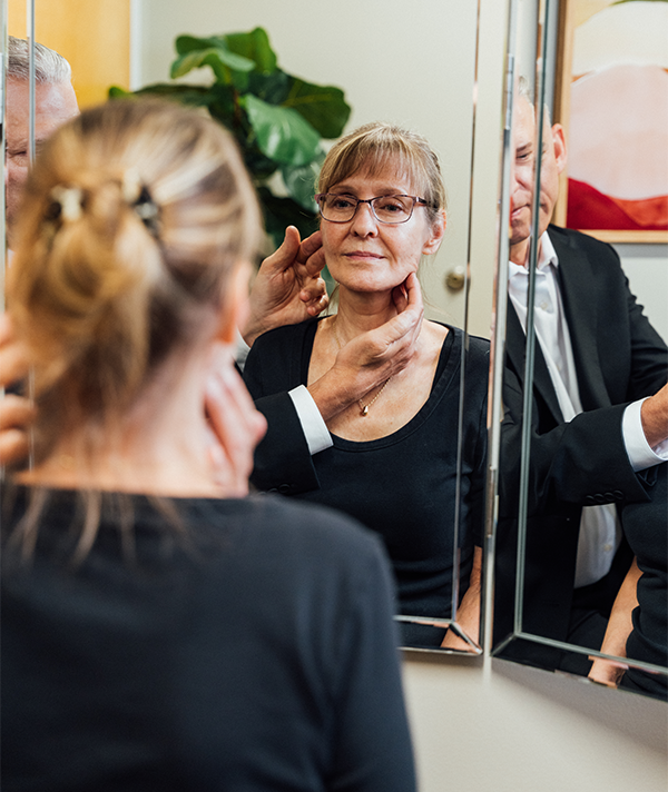 A male doctor, Dr. Lee, in a black suit, gently touching the neck area of a female patient who has her back to the camera while they both look at her reflection in a triple-panel mirror. Abstract artwork hangs on the wall behind them. - Facial Fat Grafting in Severna Park, MD
