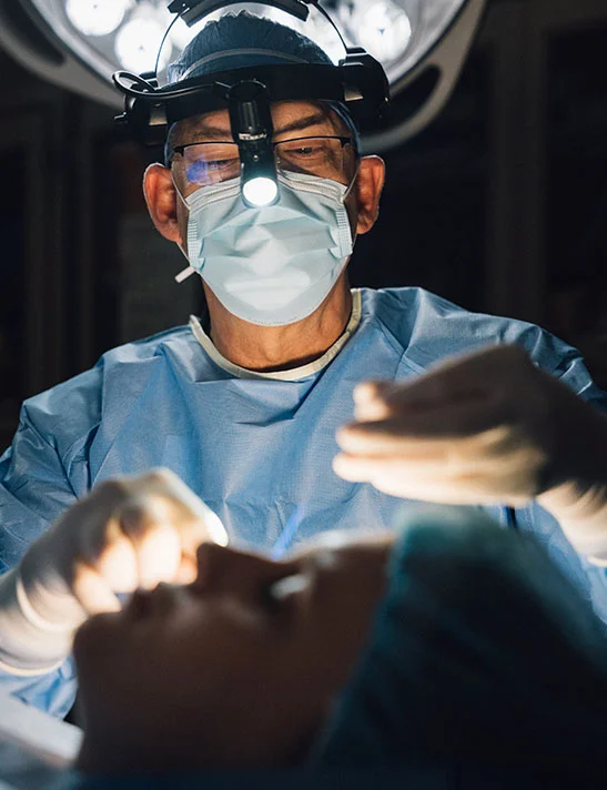 A image showing Dr. Washal, a surgeon, performing a procedure on a patient. He is wearing a surgical mask, glasses, a headlamp, and surgical gloves, with his hands near the patient's face. - Functional Rhinplasty in Severna Park, MD