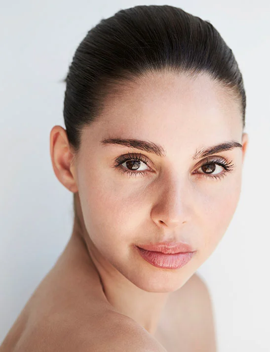 A woman with a light complexion poses against a bright white backdrop, her hair pulled back in a bun. Her gaze is calm and direct. Her natural skin texture and features, creating an impression of beauty and well-being. - Lip Augmentation Severna Park, MD