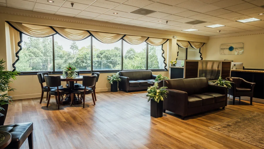 A view of a bright, contemporary medical office waiting area featuring hardwood floors, large windows with cream-colored valances, dark leather sofas, and a round table with chairs. Potted plants and a reception desk are also visible in the room.