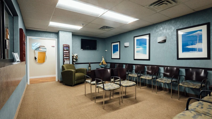 A view of a medical office waiting room with light blue textured walls, tan carpet, and fluorescent lighting. The room contains rows of dark leather and patterned chairs, a green upholstered armchair, a television, and framed blue-themed artwork.
