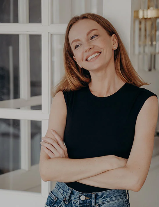 A smiling, attractive woman with light brown hair, wearing a black sleeveless top and blue jeans, standing indoors with her arms crossed, leaning against a white framed door or window. - Lower Eyelid Surgery in Severna Park, MD