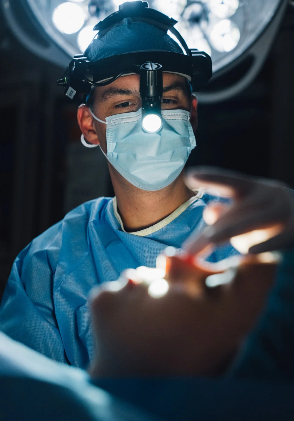 A close-up of Dr. Washal, a surgeon, wearing light blue scrubs, a surgical cap, and a mask, focused intently while performing a procedure. He is wearing a headlamp that casts a bright light onto the patient in the foreground.