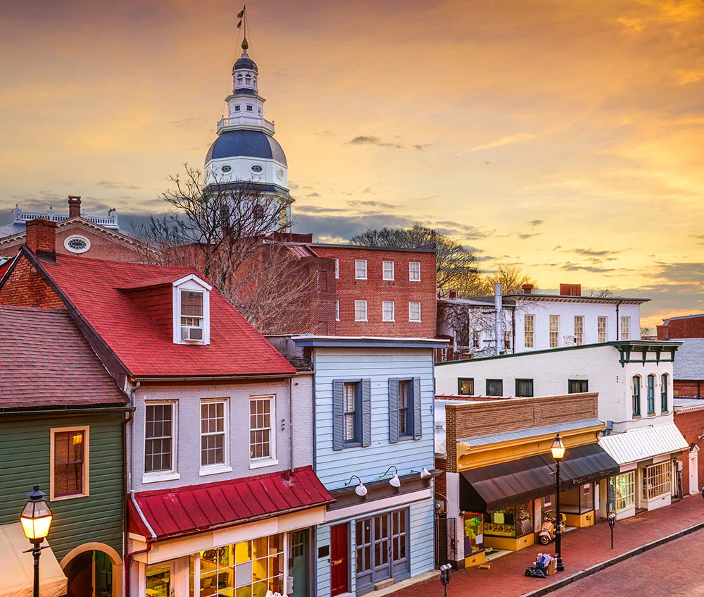 A scenic view of historic downtown Annapolis, Maryland, at sunset, showing colorful storefronts along a brick street with the dome of the Maryland State House rising in the background. - Out of Town in Severna Park, MD