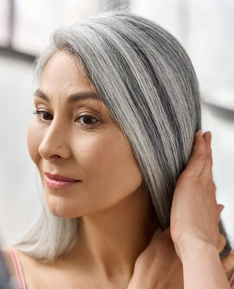 A close-up portrait of an elegant woman with long, straight silver-gray hair and smooth skin, looking to the side and gently touching her hair with her right hand.