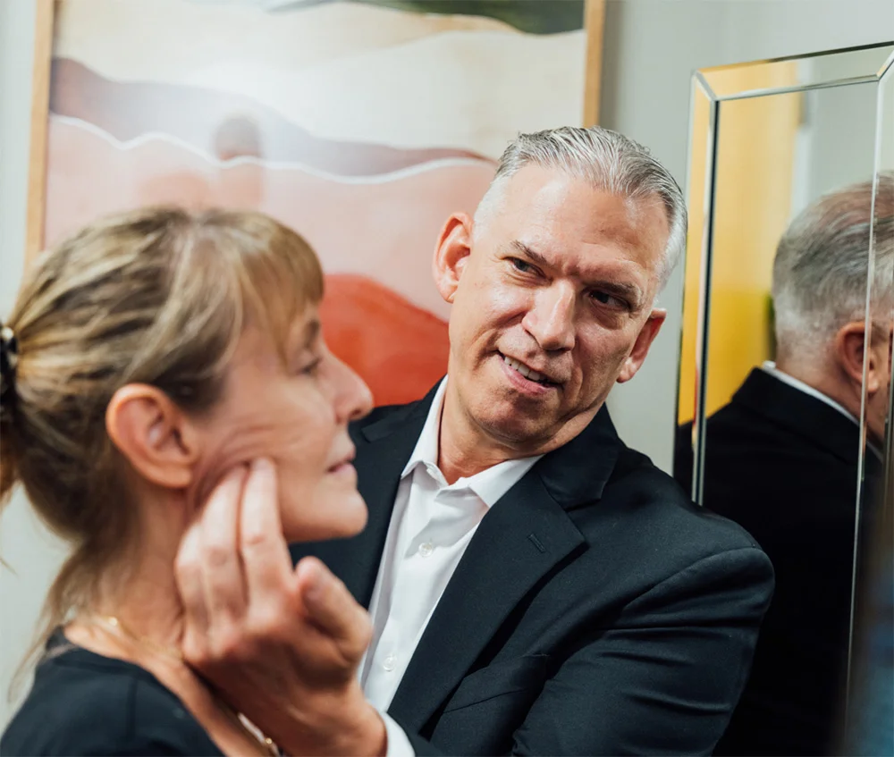Dr. Lee in a black suit and white shirt consulting with a female patient. The patient is touching her cheek while the doctor looks toward the camera with a slight smile. A mirror and abstract artwork are visible in the background.
