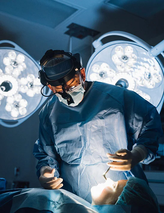 A surgeon, Dr. Lee, in blue scrubs, a surgical mask, and a headlamp with magnifying loupes, performing a procedure on a patient in a brightly lit operating room.- Revision Rhinoplasty in Severna Park, MD