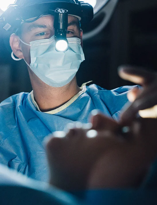 A close-up of Dr. Washal, a surgeon, wearing light blue scrubs, a surgical cap, and a mask, focused intently while performing a procedure. He is wearing a headlamp that casts a bright light onto the patient in the foreground. - Rhinoplasty in Severna Park, MD