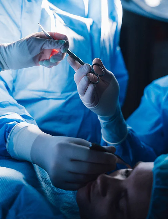 A close-up image inside an operating room showing gloved hands in blue surgical gowns. One hand holds scissors, transferring a surgical tool to the other hand above a patient's face during a procedure. - Scar Revision in Severna Park, MD
