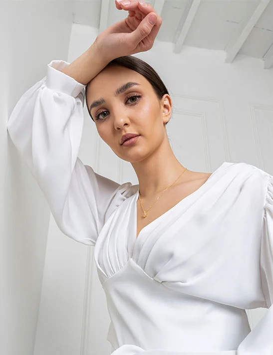 A close-up, low-angle portrait of a woman with dark hair pulled back, wearing a white v-neck top with puffed sleeves and a delicate gold necklace. She is leaning against a white wall with her right arm raised above her head, looking directly at the camera. - Upper Eyelid Surgery in Severna Park, MD